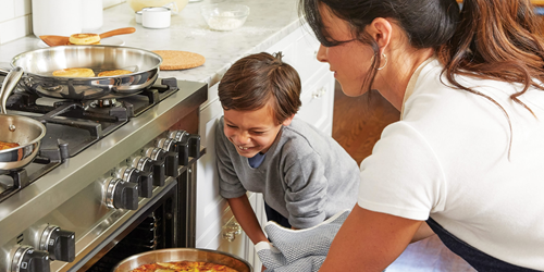 A woman putting a pan in the oven with a young child nearby watching and smiling.